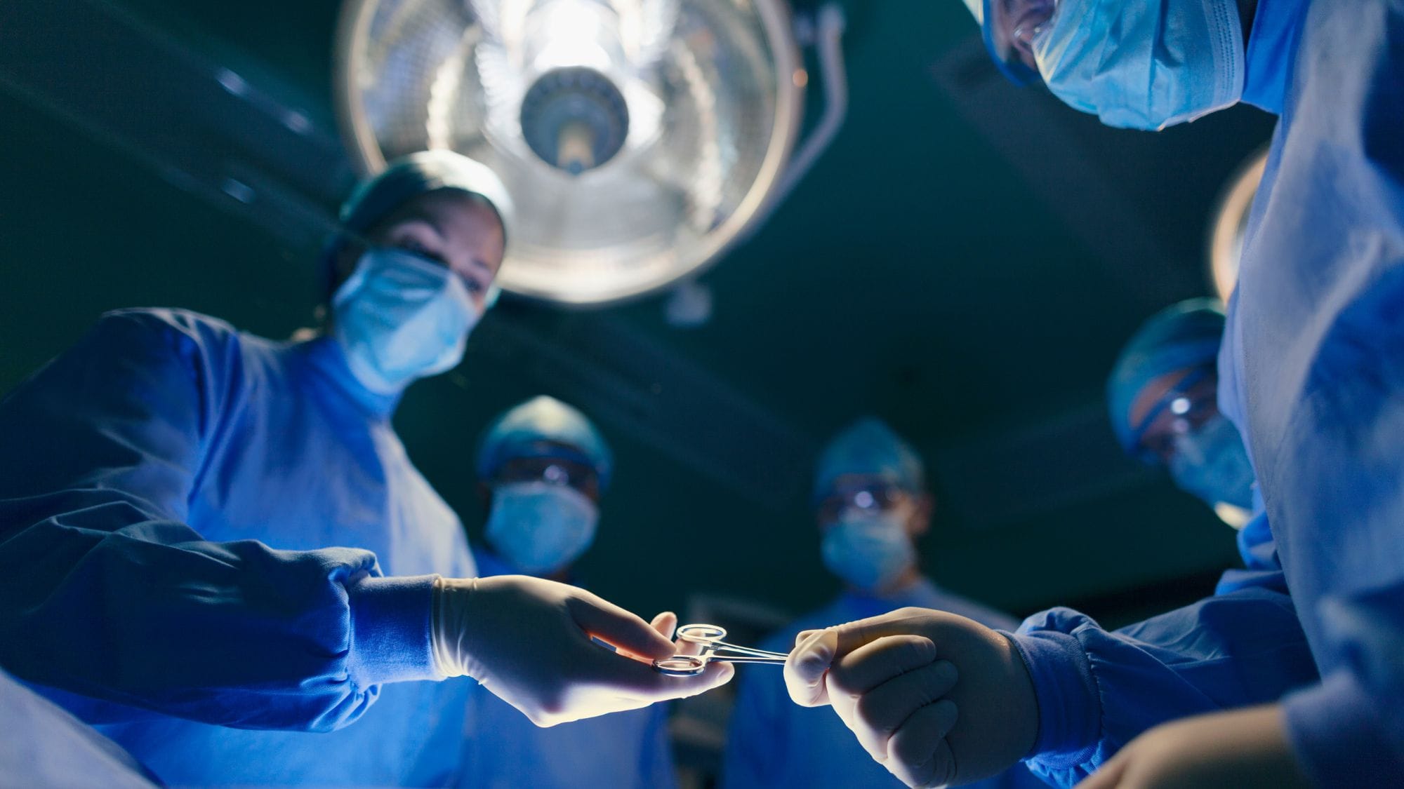 Green operating room with doctors, nurses, and other medical personnel dressed in blue.